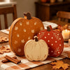 A set of three wooden pumpkin figurines in orange, red, and cream are displayed on a checkered cloth with autumn leaves.