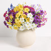 A vibrant bouquet of artificial silk flowers in yellow, purple, and white arranged in a cream-colored vase.