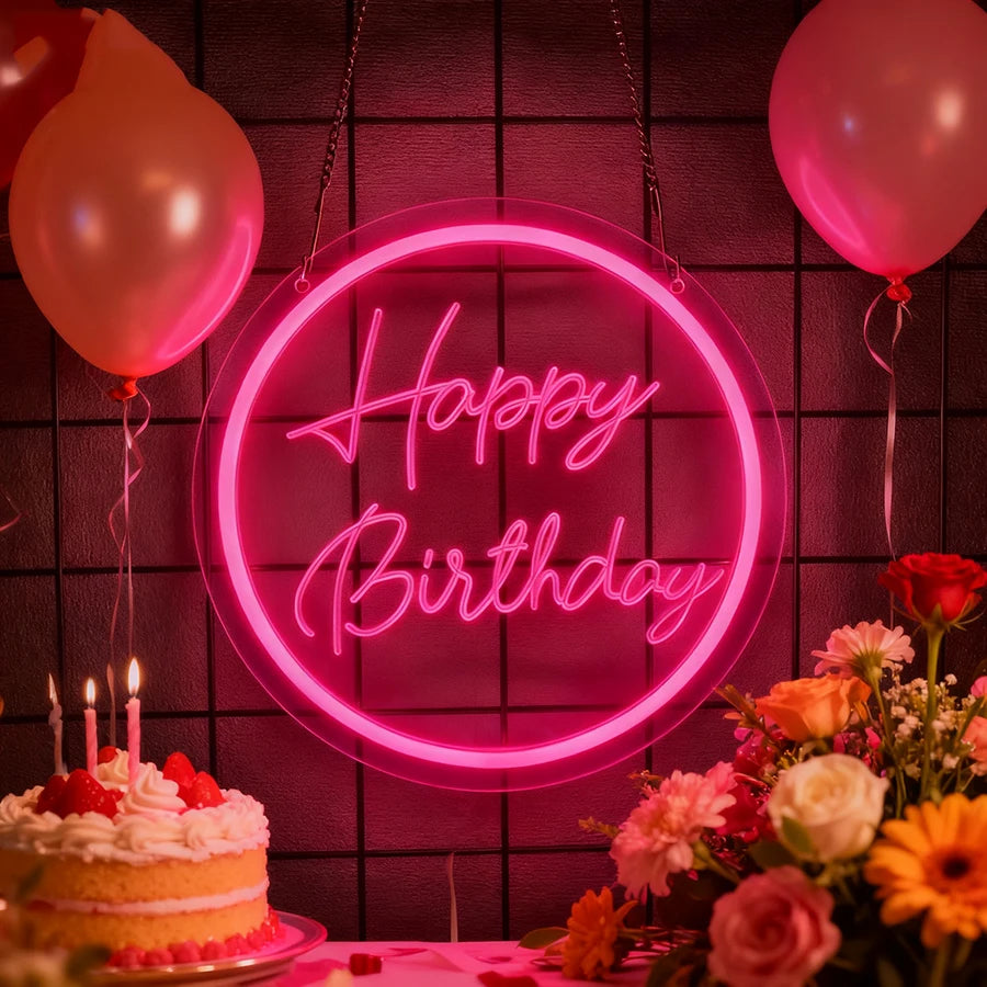 A pink neon sign shaped like a birthday message hangs against a dark brick wall with a cake and flowers below.