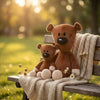 A brown teddy bear sits on a wooden bench outdoors with soft sunlight and a knitted blanket.