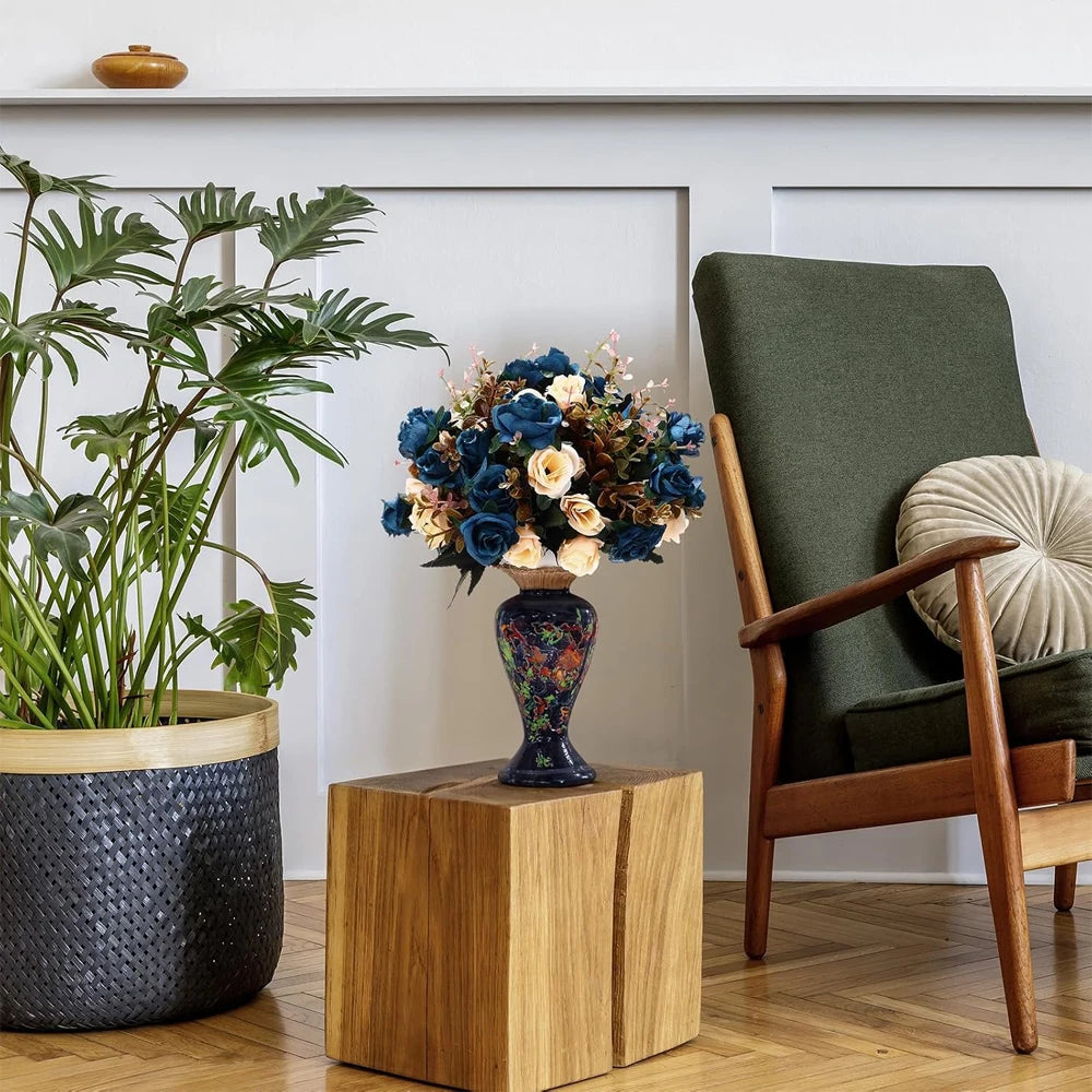 A vase of artificial silk roses in blue and cream colors sits on a wooden block next to a green armchair.