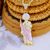 A gold-plated pendant necklace featuring a figure of Saint Jude surrounded by a pink drape, resting on woven mats.