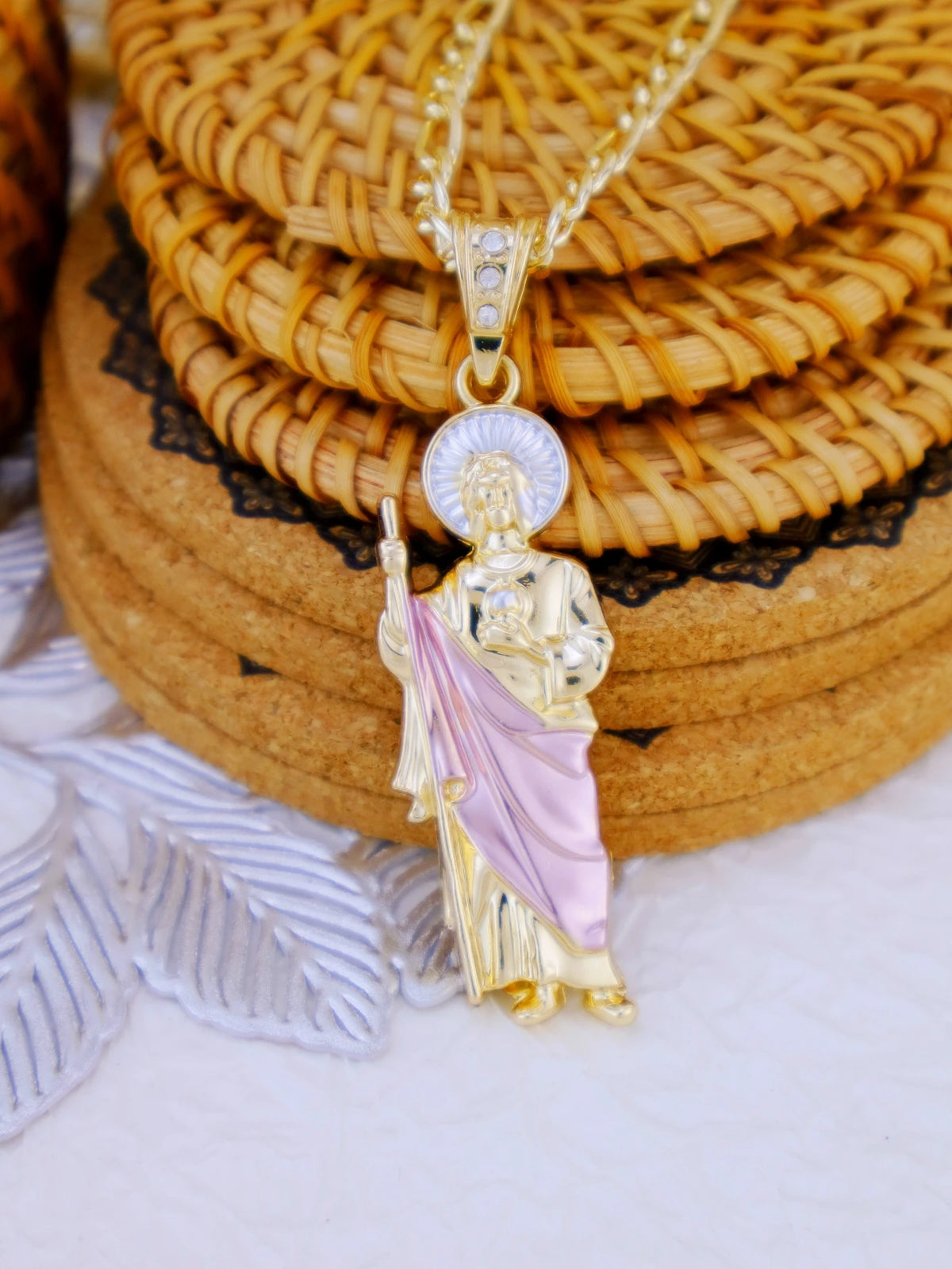 A gold-plated pendant necklace featuring a figure of Saint Jude surrounded by a pink drape, resting on woven mats.