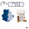 A blue and white knitted plush shark character with an emotional support tag next to a beige cup plush.