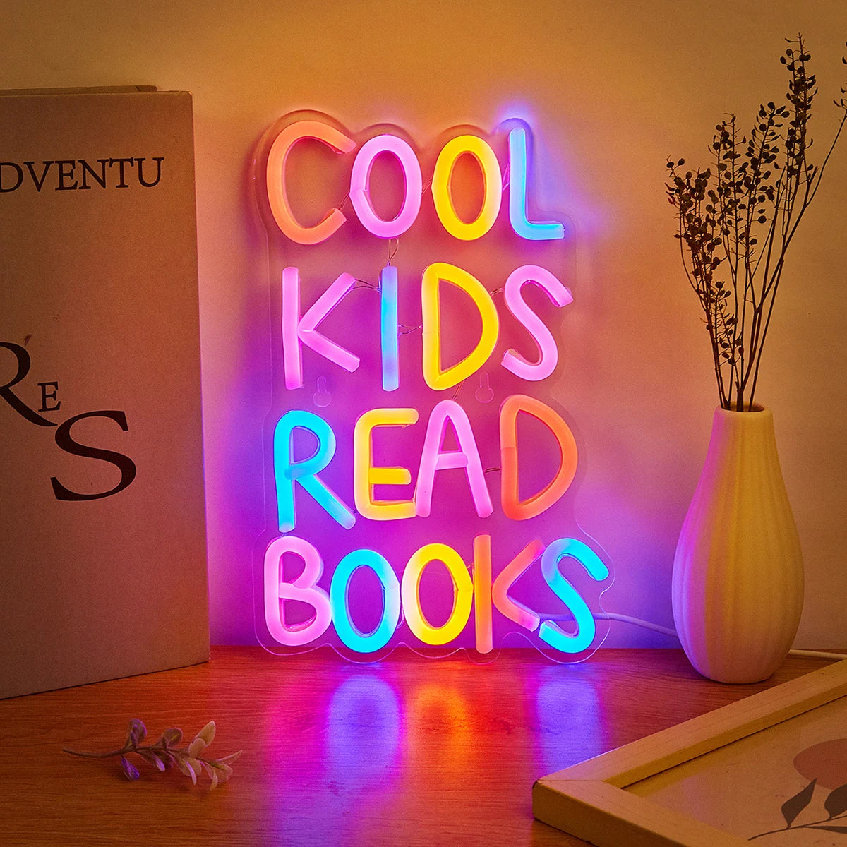 A neon sign spelling out COOL KIDS READ BOOKS illuminated on a wall next to a vase of dried branches.