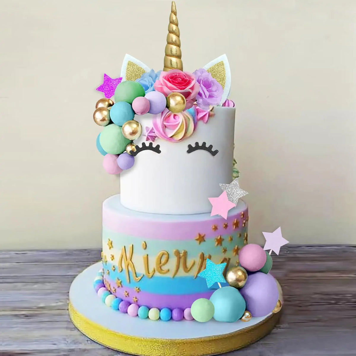 A tiered white cake decorated with pink roses, colorful balls, and a golden unicorn horn.