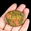 A hand holds a smooth, oval green stone with orange inclusions engraved with the word Lucky.