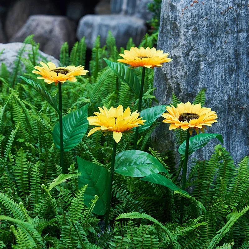 Yellow solar sunflower LED garden lights with three adjustable heads growing among green ferns and rocks.