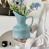 A hand holds a modern blue ceramic tabletop vase filled with blue and white flowers.