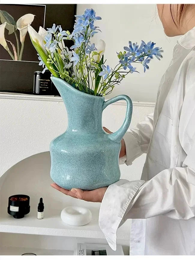 A hand holds a modern blue ceramic tabletop vase filled with blue and white flowers.