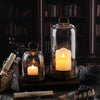 Antique brass metal candle holders with glass domes displaying glowing candles on a dark wooden surface.