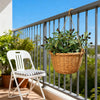 An artificial olive tree branch with green leaves and dark berries hangs in a woven basket on a balcony railing.