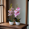A realistic artificial orchid potted plant with pink and purple flowers displayed on a wooden surface.