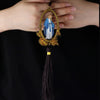 A wooden pendant ornament depicting Our Lady of Grace with a blue and white robe, held by a person's hand.