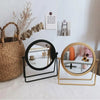 A set of two circular metal makeup mirrors, one black and one gold, displayed on a white surface.