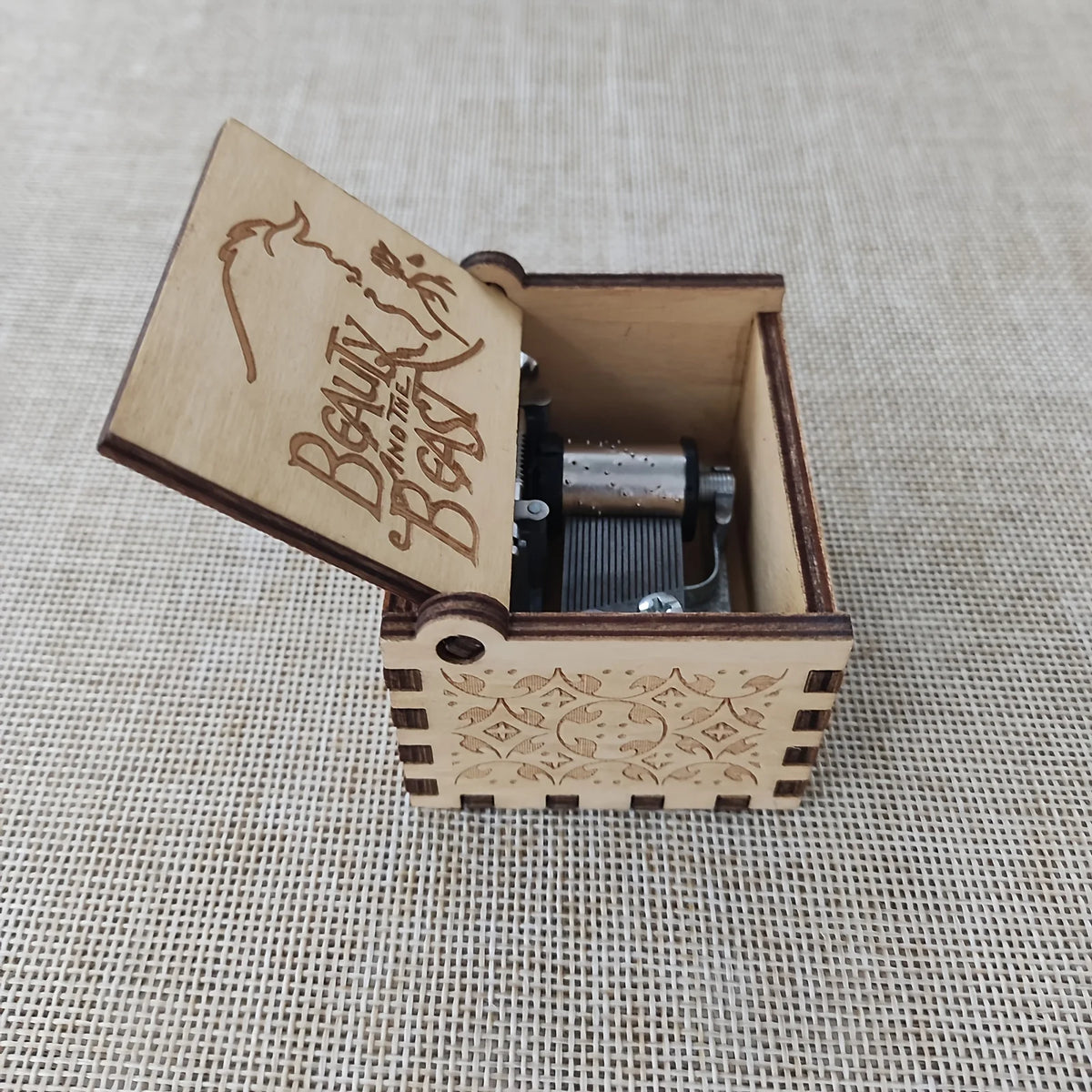 An antique carved wooden music box with a lid displaying the text Beauty and the Beast resting on a woven background.
