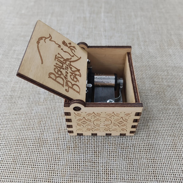 An antique carved wooden music box with a lid displaying the text Beauty and the Beast resting on a woven background.