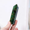 A hand holds a vibrant green emerald crystal point healing wand with natural veining against a white background.