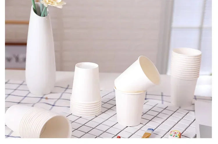 A collection of white disposable paper cups arranged on a checkered surface with a vase of flowers in the background.