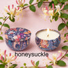 A jar of soy wax candle with a floral pattern sits next to its lid on a pink background surrounded by greenery and flowers.