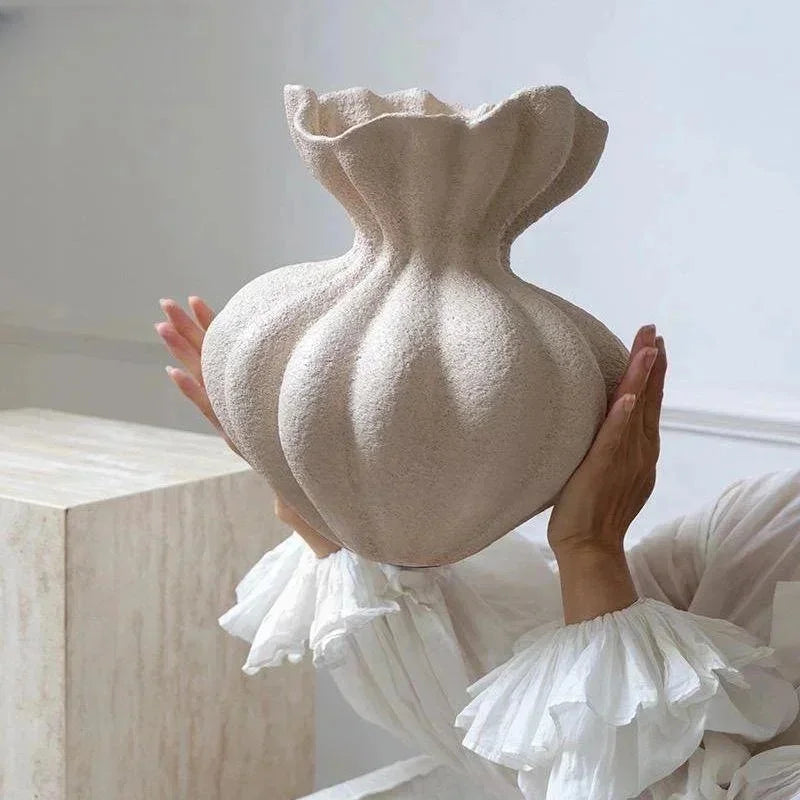 A hand holds a beige, organic-shaped ceramic vase with ruffled white fabric in a bright room.