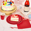 A red paper party tableware set featuring dinner plates, napkins, cups, and cutlery is displayed on a white tablecloth.