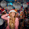 A smiling woman wearing a pink blazer and gold garland is surrounded by silver disco ball foil balloons in a party setting.