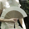 A person holds a classic bamboo folding fan with a decorative pendant against a natural background.