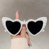 A hand holds a pair of heart-shaped glasses decorated with white pearls and dark lenses.