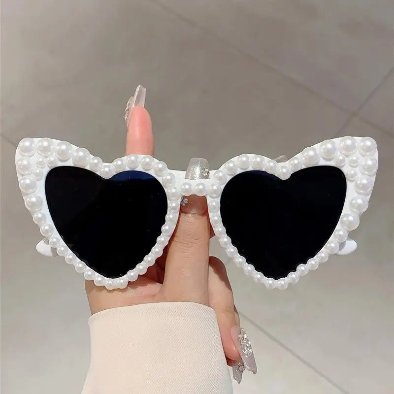 A hand holds a pair of heart-shaped glasses decorated with white pearls and dark lenses.