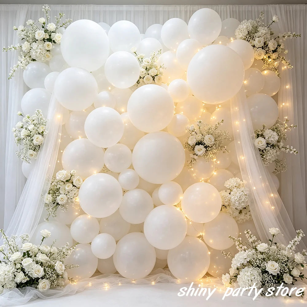 A large arrangement of matte white latex balloons decorated with white flowers and illuminated by string lights.