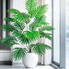 A vibrant green artificial palm tree with silk leaves displayed in a white pot next to a window.