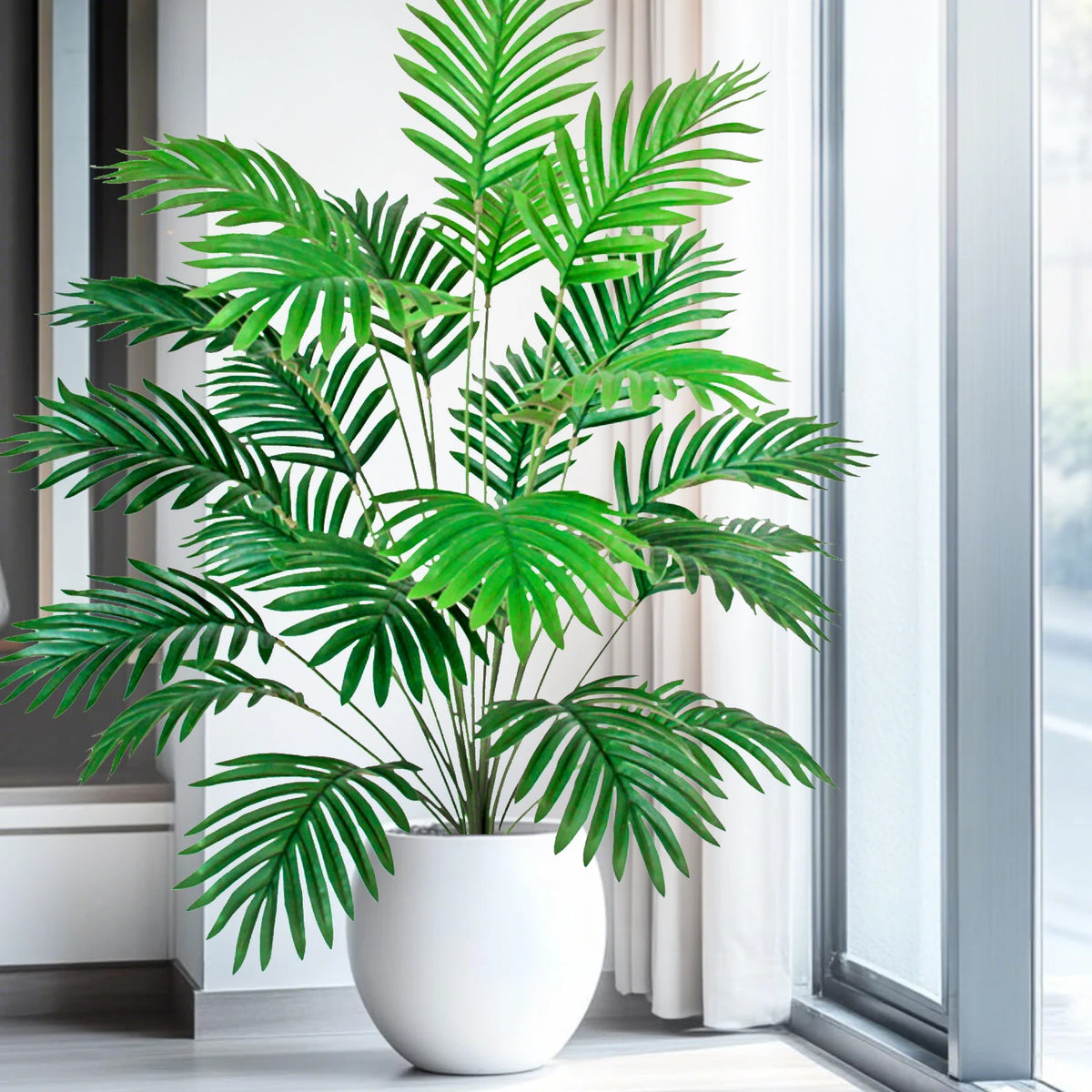 A vibrant green artificial palm tree with silk leaves displayed in a white pot next to a window.