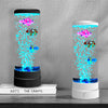 A tall glass lamp displays colorful, glowing fish swimming inside, set on a wooden surface.