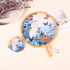 A circular fabric hand fan featuring a blue and white bird design with butterflies, shown next to a folded version.