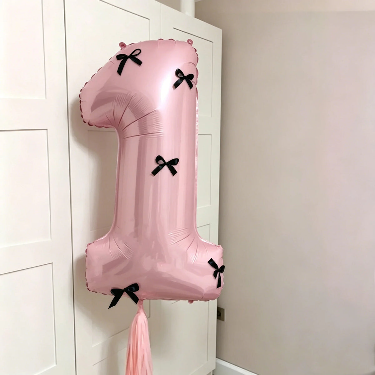 A large pink number balloon shaped like '1' with black bows, displayed next to a white cabinet.