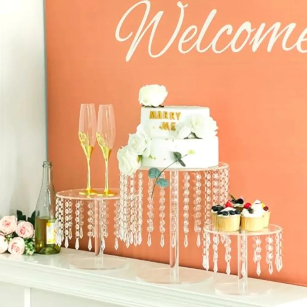 A white wedding cake with 'Marry Me' inscription displayed on a clear acrylic stand with dangling crystals.