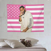 Un homme en chemise claire se tient devant une tapisserie de drapeau américain rayée rose et blanc.