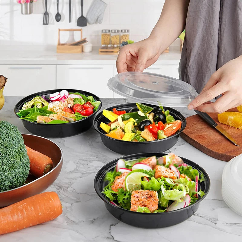 A hand is preparing a meal in black round containers with clear lids, surrounded by fresh vegetables and citrus fruit.