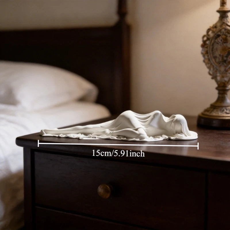 A white draped sculpture of a reclining female figure is placed on a dark wooden surface.