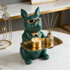 A bronze green bulldog figurine sitting on a gold tray with a lit candle and surrounded by decorative gold accents.