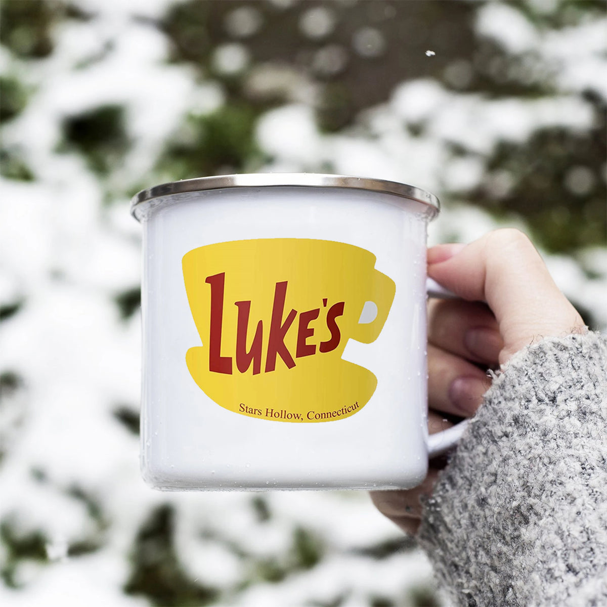 A white enamel coffee mug featuring a yellow silhouette of a lion's head with the text Luke's on it, held against a snowy out