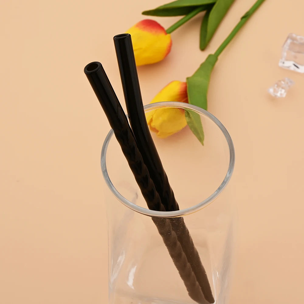 Black reusable borosilicate glass straws with a cleaning brush displayed in a glass container next to yellow tulips.