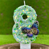 A handmade candle shaped like the number 8 covered in green sequins with a butterfly decoration.
