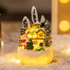 A glowing resin ornament shaped like a snowy gingerbread house with evergreen trees and festive figures.