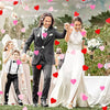 A happy bride and groom in wedding attire surrounded by falling pink and red heart confetti outdoors.