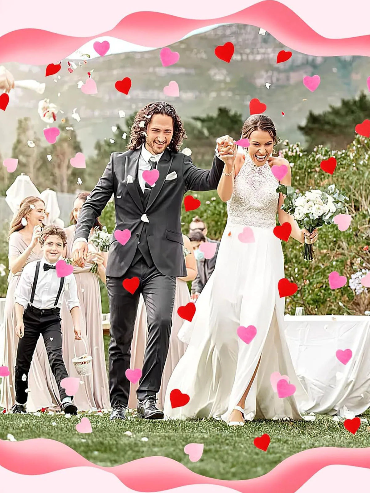 A happy bride and groom in wedding attire surrounded by falling pink and red heart confetti outdoors.