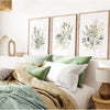 Three framed watercolor botanical prints featuring green and white wildflowers displayed on a bed.