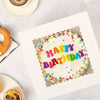 A white birthday cake decorated with colorful sprinkles and the words HAPPY BIRTHDAY, presented in a white bakery box.