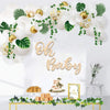 A wooden sign spelling out Oh Baby decorated with white balloons and green foliage for a baby shower